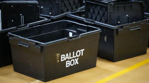 A collection of big black plastic ballot boxes, with one reading ballot box in white writing