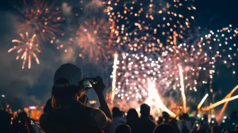 A stock image the back of a girl in a baseball cap filming a fireworks display on her mobile phone.