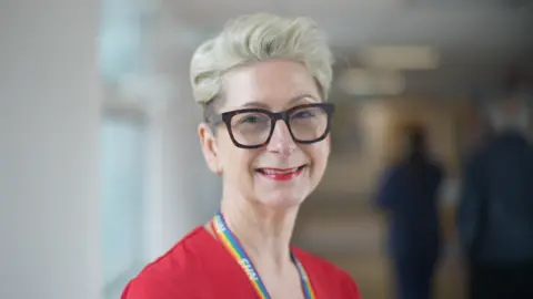 Nicola Cottington with short light coloured hair, wearing glasses and a red top and a rainbow NHS lanyard around her neck.
