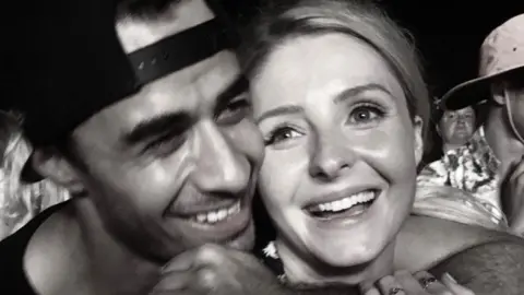 Kelsey Mulcahy A black and white image of a man and a woman in a crowd. The man is wearing a backwards cap and is smiling, with his arm around a blonde woman who is also smiling.