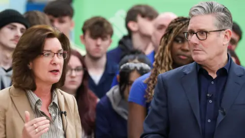 Eluned Morgan, a woman with brown hair and glasses, speaks in front of a crowd of people while standing next to Sir Keir Starmer, a man with grey hair and glasses.