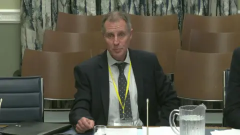 NI Assembly Micheál Mac Giolla Ghunna speaking at the Stormont Education Committee. He is wearing a suit, a polka dot tie, white shirt and grey hair.