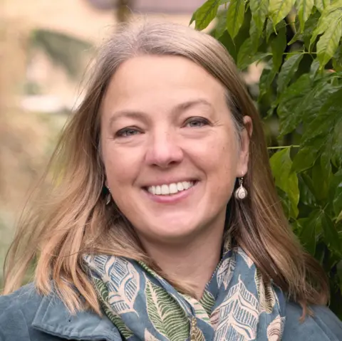 RSPB Katie-Jo Luxton, wearing a green coat and patterned scarf, is standing in front of a leafy background and smiling.