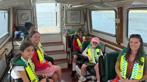 Four small children with life jacked on sit on a boat.