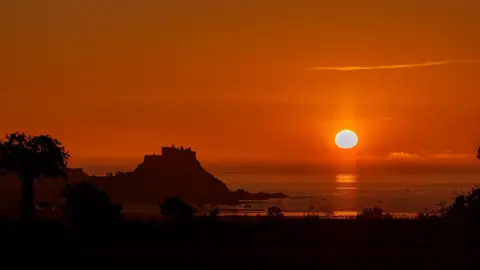 BBCWeatherWatcher/Daniel Rive The sun is bright orange as it rises over and is reflected in the sea. The sky is dark orange and a castle on an island is starkly black in silhouette. 