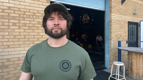 Arzu Dutta/BBC Annie's coach, Luke Stopford, is standing outside his gym. Luke is wearing a khaki green top and wearing a matching sports cap. 