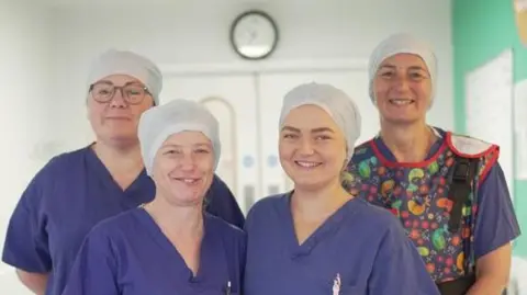 Four nurses in uniform. They are stood in a corridor and smiling at a camera. The walls around them are white with display boards on either side. Behind them are white doors and a clock. 