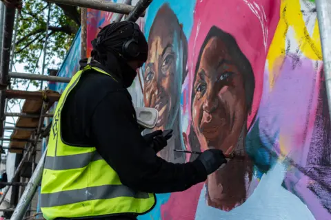 Toyin Adedokun / AFP via Getty Images A man in a high-vis jacket paints a mural of a woman. He is surrounded by scaffolding.