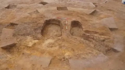 OCA An archaeological site showing sandy soil and two excavated graves in the middle of a barrow - the one on the left much larger than the one on the right, which is slightly lower down the barrow. 