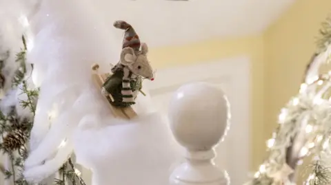 Kirsty Anderson Stuffed mouse figurine wearing a striped hat and scarf, skiing down a banister covered in fake snow and Christmas lights