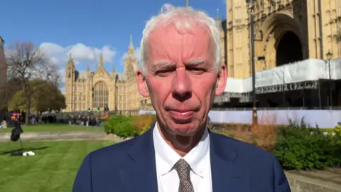 An elderly man in a suit. He is standing outside the Houses of Parliament. 