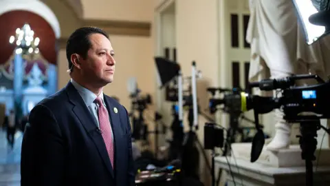 Tony Gonzales stands in a navy suit, blue shirt and red tie while looking down the lens of a television camera. He is standing in a regal-looking hallway where other television cameras are lined up. 