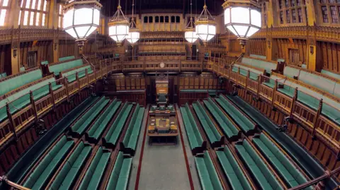 The Commons Chamber, a wood-panelled room of rows of green benches facing each other, with a space of floor and a large table in the middle. There is a gallery above the benches with three rows of green benches on it. A large chair at one end of the room. Windows high up in the room and six lights hang on pendants from the ceiling.
