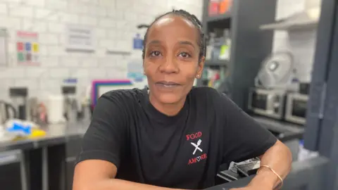 A woman wearing a black shirt looks at the camera while standing in a commercial kitchen.