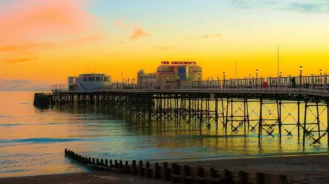Getty Images A pier stretching out into the sea is bathed in orange and yellow light, with calm water around it. A building in the middle of the pier has a large sign on top saying "pier amusements".