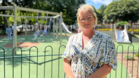 Sonja is in a floral dress stands behind a black metal fence in front of a playground with swings and slides. Children are playing in the background, and trees and a clear blue sky are visible