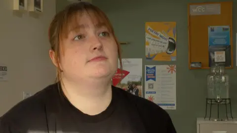A woman in a black t-shirt speaks in profile to someone unseen. She has light red hair, freckles and a nose ring. Behind her is a cream and green wall with flyers stuck to it.
