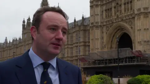 Winchester MP Danny Chambers standing in front of the Houses of Parliament wearing a blue suit with a blue tie.