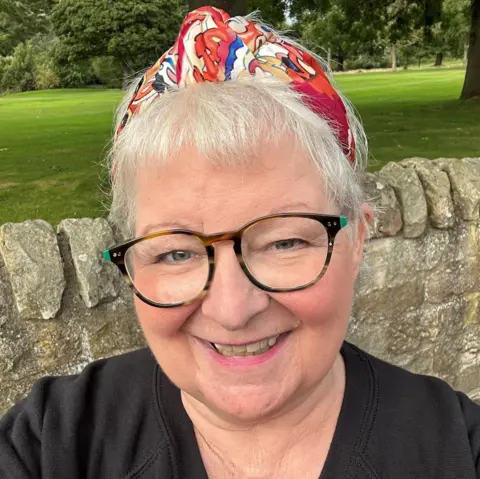 PA Media Janey Godley, with abbreviated  grey hair, acheronian  glasses and a reddish  bandana connected  her head, smiles astatine  the camera with a chromatic  partition  and greenish  parkland  down  her 