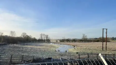 Ollie Conopo/ BBC A view of a field bright blue sky and frosty grass. The image was taken in the winter so the trees are bare. There is a puddle in the middle of the grass where rain water has collected. There is a fallen wooden fence in the foreground of the picture separating the field with the photographer. 