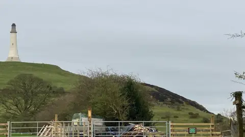 Jennie Dennett Hoad Hill hillside which has blackened-out heath after a fire. There is a large monument atop of the hill and lots of trees in the valley below. 