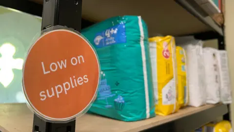 There is a shelf with some packets of nappies on with a sign saying 'Low on supplies' in a white font on an orange, circular cutout which has been laminated and attached to the shelf.