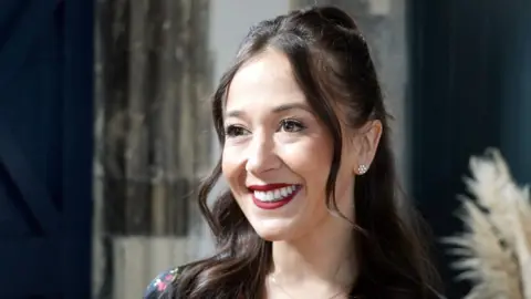 Shaun Whitmore/BBC Sophie Pinsent has long brown hair and is wearing a red lipstick. She is standing in front of a concrete wall.