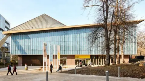 Lufton+Crow People can be seen strolling in front of a building with a sloping roof which curves upwards and pale walls. Some leafless trees can be seen to the right.
