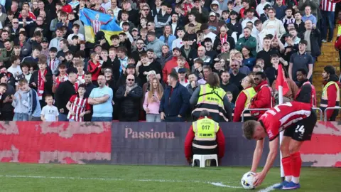 Graham Burrell A footballer placing the ball down before taking a corner during a match. There are hundreds of supporters watching from a stand, with one fan holding a Ukrainian flag aloft.