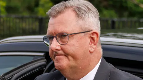 Close-up on a man's head as he emerges from a car. He has short mostly grey hair, which has a side part. He is wearing dark-rimmed glasses, a dark suit jacket and a white shirt. The car he is emerging from is dark coloured. In the background, out of focus, a metal fence and some greenery can be seen