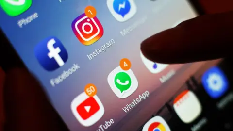 PA Media Close up image of smartphone screen showing the emblems of Facebook, Instagram, WhatsApp and YouTube, in a dark room and illuminating a person's finger hovering over the screen