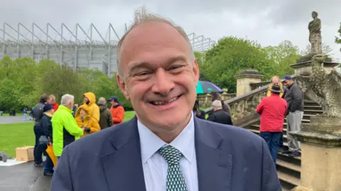 Sir Ed Davey is smiling and wearing a blue suit, patterned tie and pale shirt. There are people behind him and Newcastle United's football stadium.