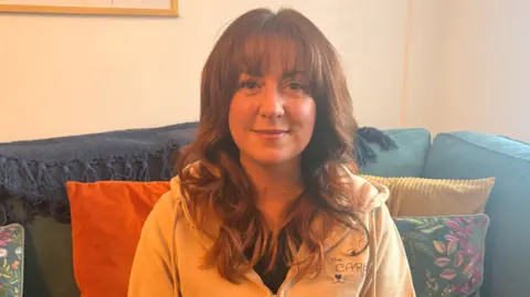Sheree had long brown hair and is wearing a black t-shirt and a cream hooded top. She has long brown wavy hair. She is sitting on a blue sofa surrounded by several cushions and is smiling at the camera.