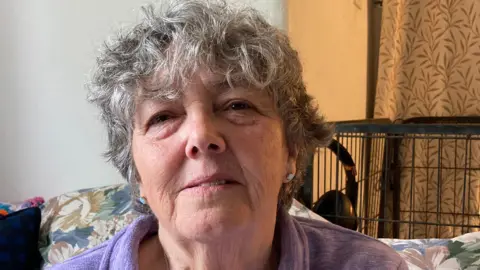 A woman with curly grey hair is looking at the camera. She is wearing a purple top, and is sitting on a floral patterned sofa.