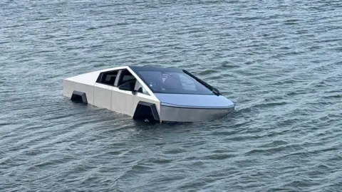 A silver Tesla Cybertruck seen partially submerged in water, which has risen half way up the wheels, in a lake.