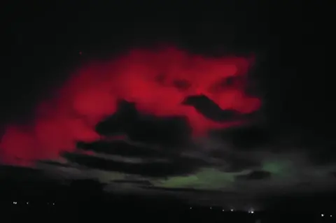 Daniel Page Pink from the northern lights shine through the clouds, the shape resembles the side profile of a face of a dragon with a grin and and an eye. Below it, the lights of the town twinkle in the night sky.