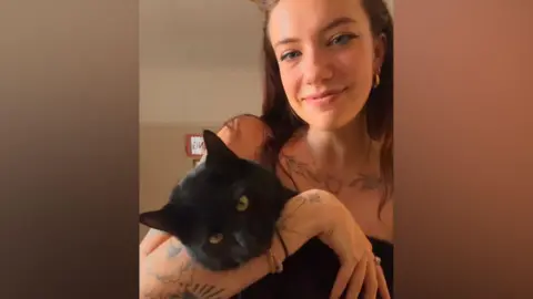 A woman with brown hair and tattoos holds a large black cat in her arms. 