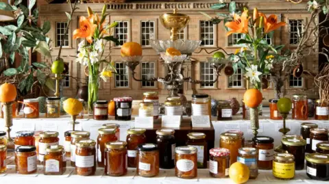Hermione McCosh Rows and rows homemade marmalade are on display with oranges on candlesticks and vivid orange lillies in vases. There appears to be a model of a stately home in the background. 