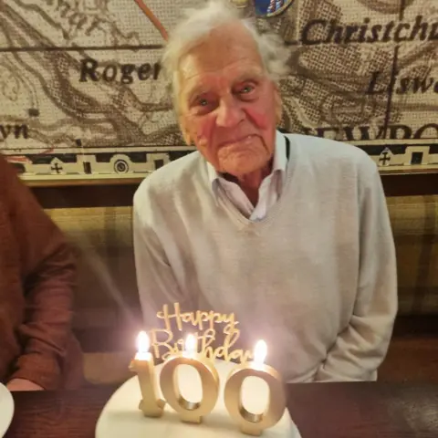 PA Media A man smiling at the camera. There is a cake in front of him with candles that say Happy Birthday 100.