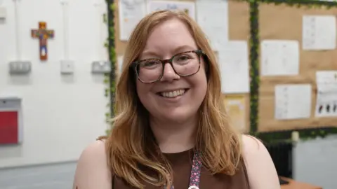Sarah Horspole standing in a primary school classroom. She is wearing a brown sleeveless top and is looking directly at the camera and smiling.