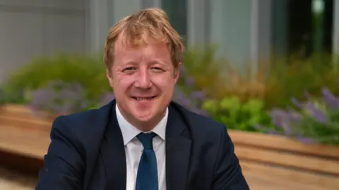 BBC Paul Bristow, wearing a black suit and a white shirt and a navy tie, sits on a bench and smiles for the camera. 
