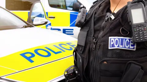 The images shows the front of a police car and an office standing next to it, with a strip saying "police" on the right-hand-side behind a hand-held radio. There is also another police car behind the police car.