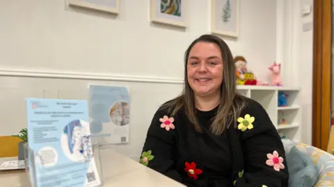 Cat smiles at the camera as she's sat at a table. She is wearing a black jumpers with knitted flowers on it. She has light brown hair and behind is shelving with cuddly toys on it.