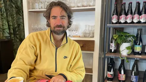 Bianca Ramsey A man with wavy brown hair and a beard sits indoors wearing a yellow Nike fleece zip-up jacket. In the background, white shelves display glassware, and a black metal rack holds wine and beer bottles alongside a small potted plant.