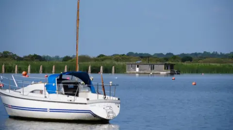 Shaun Whitmore/BBC A view of Oulton Broad. A yacht rests in the water with a houseboat in the distance.