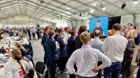 Journalists, including the BBC's political editor Chris Mason, watch a screen showing Ukrainian leader Volodymyr Zelenskyy