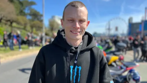 Stuart Woodward/BBC Danny Clipson wears a black hoodie with a blue Monster logo on the chest. He also has a black lanyard around his neck. He has cropped brown hair and is smiling at the camera. There are motorbikes blurred out behind him and a ferris wheel.