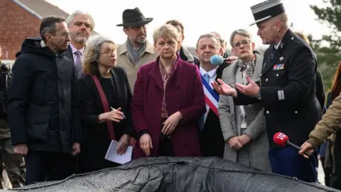 Yvette Cooper wearing a purple coat, centre, looks towards the camera at a black dingy. On the left, Bruno Retailleau looks past her to the right at a member of the army in a black coat who speaks to them both. Behind and around them are various other officials and members of the media