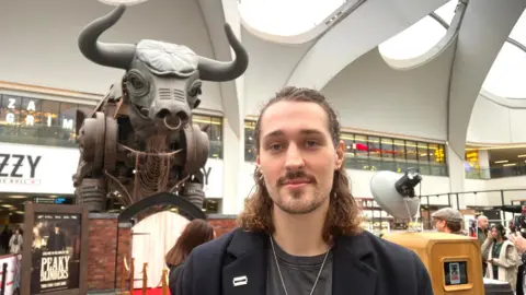 A man with curly brown hair looking into the camera with a small smile, he has a beard and a nose piercing. Behind him is a mechanical bull which has a flat cap on it's head. The flat cap is grey.