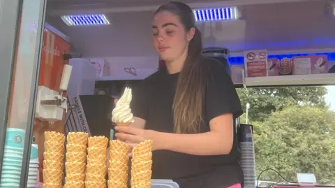 A woman is scooping soft serve onto a waffle cone. She has long hair and is wearing a black shirt.